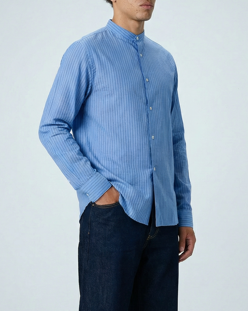 Liam band collar shirt in light blue and blue stripe, worn untucked in full-length side angle view with hand in pocket, showing relaxed fit and crinkle cotton voile fabric.